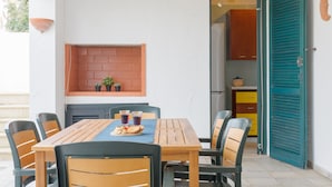 Un coin repas avec une table et des chaises en bois, une étagère en briques ornée de plantes, et une cuisine visible à travers une porte ouverte.