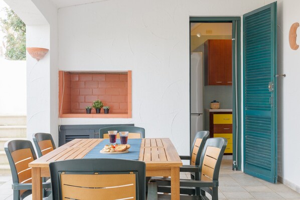 A dining area with a wooden table and chairs, a brick wall shelf with plants, and a kitchen visible through an open door.
