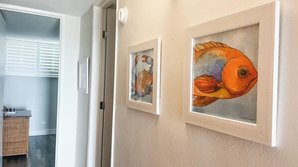 A hallway with two framed paintings of fish on the wall, a door leading to a room with a wicker cabinet, and a window with blinds.