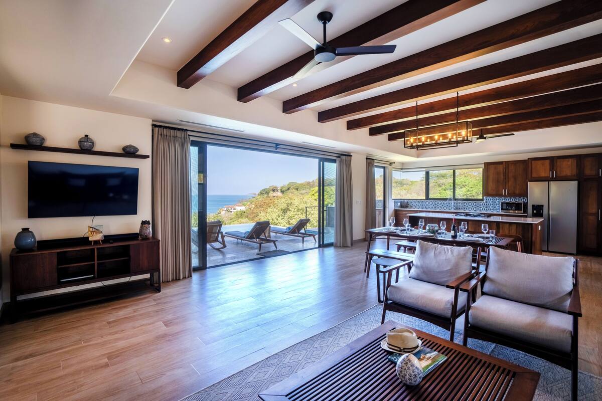 A modern living room with a large TV, wooden ceiling beams, a dining area with a view, and a kitchen with wooden cabinets.