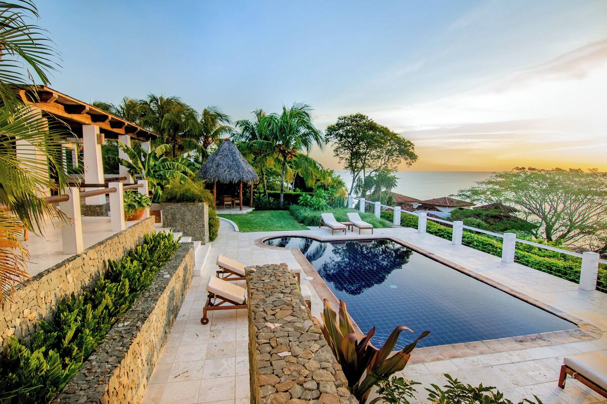 A pool area with lounge chairs, a stone wall, and a view of the ocean at sunset.