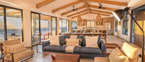 Un vaste espace de vie comprenant un grand canapé, une table basse et des fauteuils. On y trouve également des ventilateurs de plafond, un coin repas avec une cuisine en arrière-plan, et de grandes fenêtres offrant une vue dégagée.