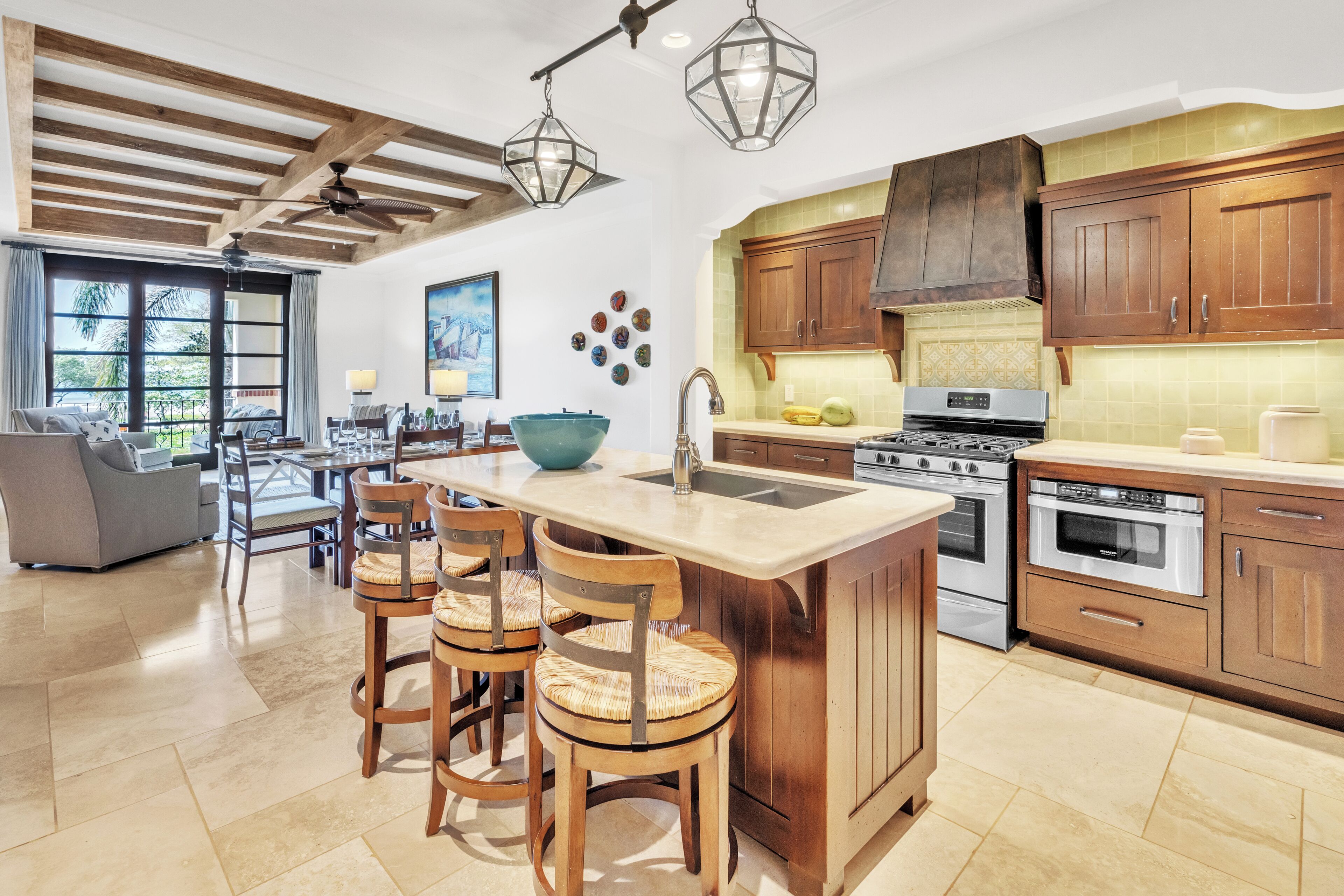 A modern kitchen with wooden cabinets, a large island with bar stools, and a dining area with a gray sofa and chairs.