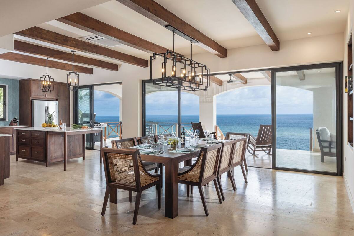 A spacious dining area with a large table, wooden chairs, and a view of the ocean.