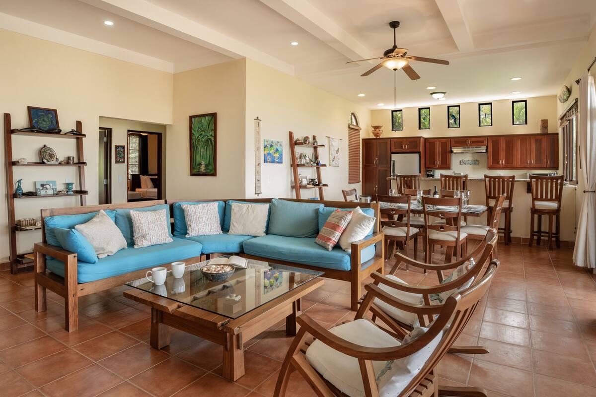 A spacious living area with a blue sectional sofa, wooden chairs, a glass coffee table, and a dining area with wooden chairs and a table.