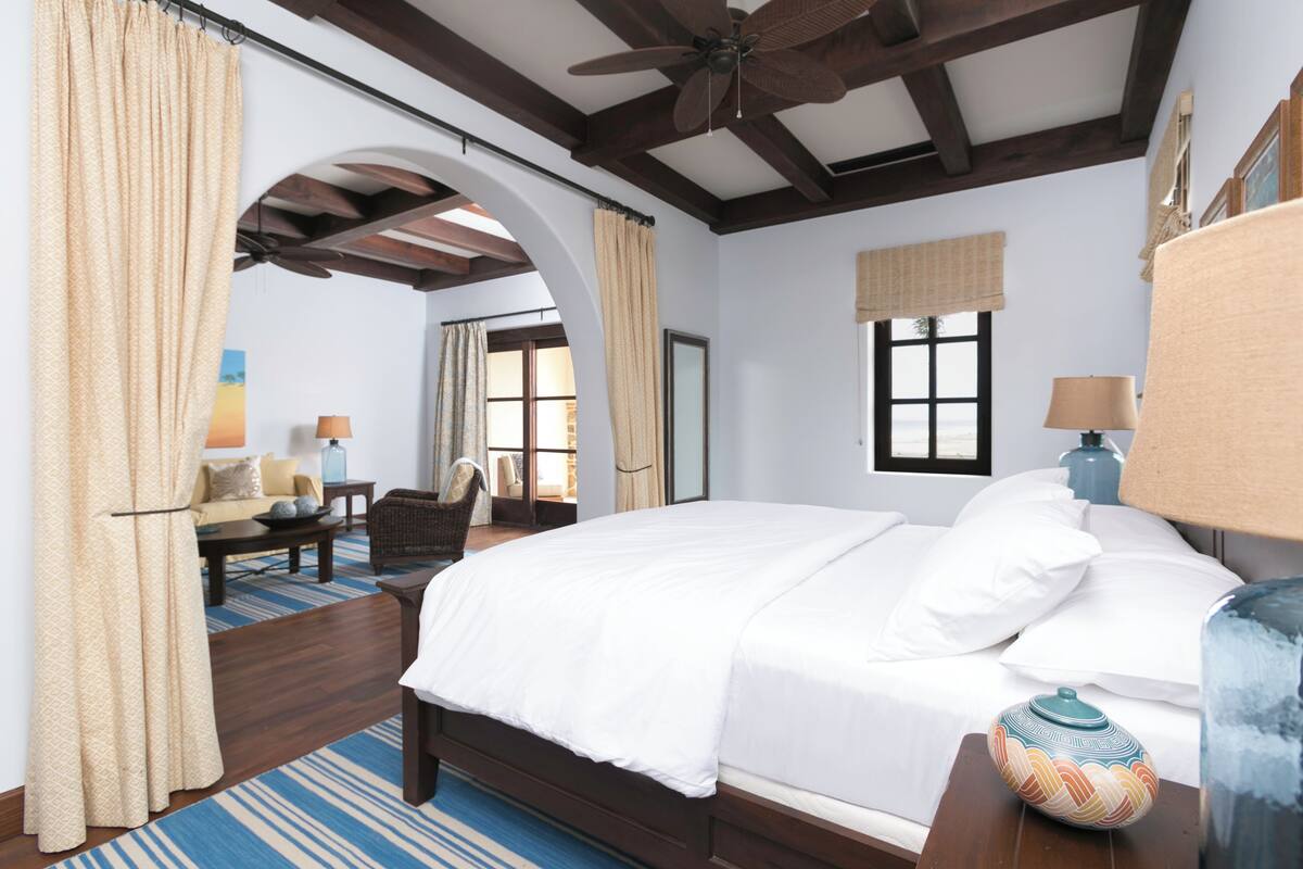 A bedroom with a large bed, a ceiling fan, a window with curtains, a lamp, and a vase on the nightstand.