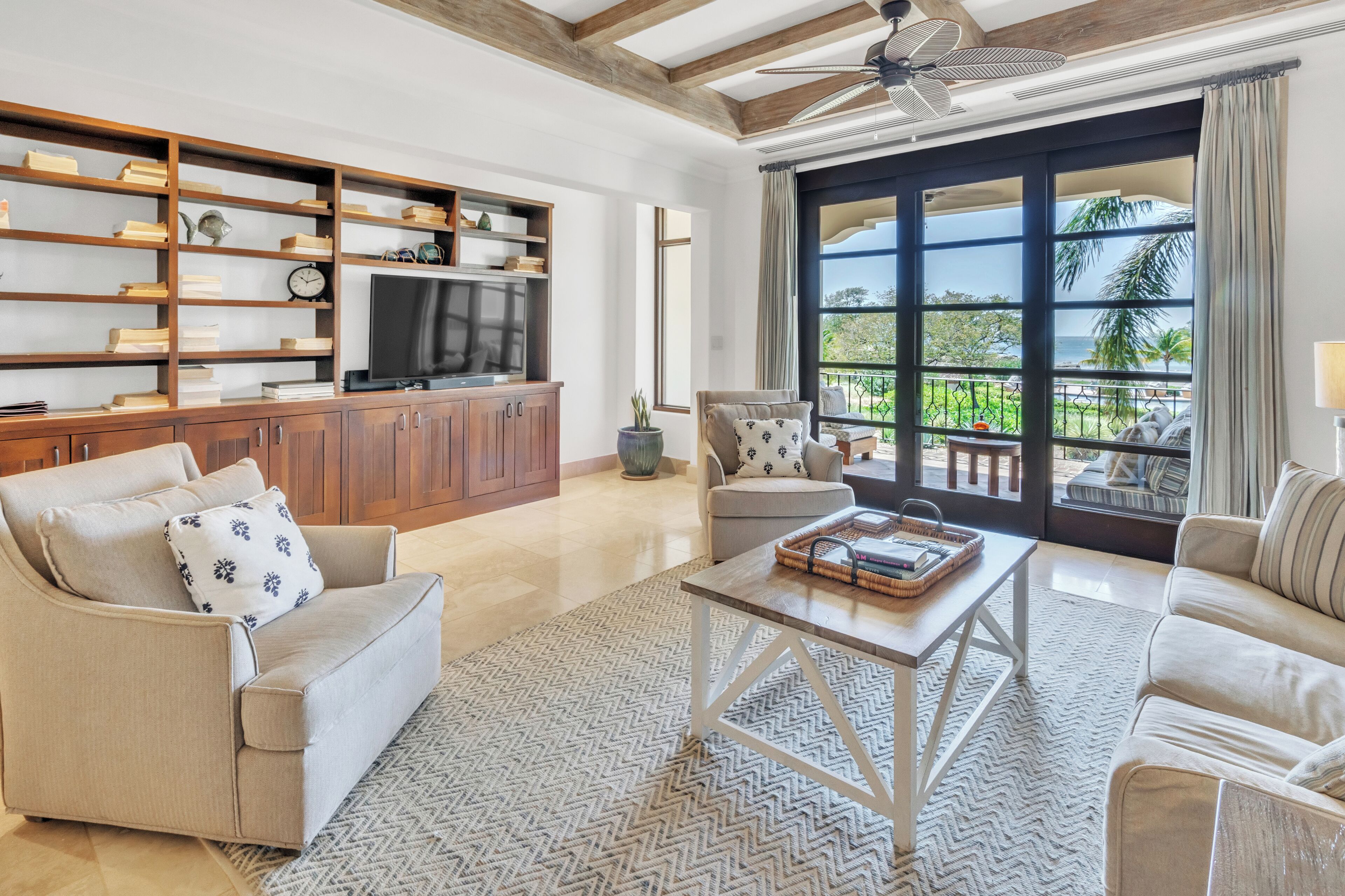 A spacious living room with a wooden bookshelf, a television, and a large window with a view of palm trees.