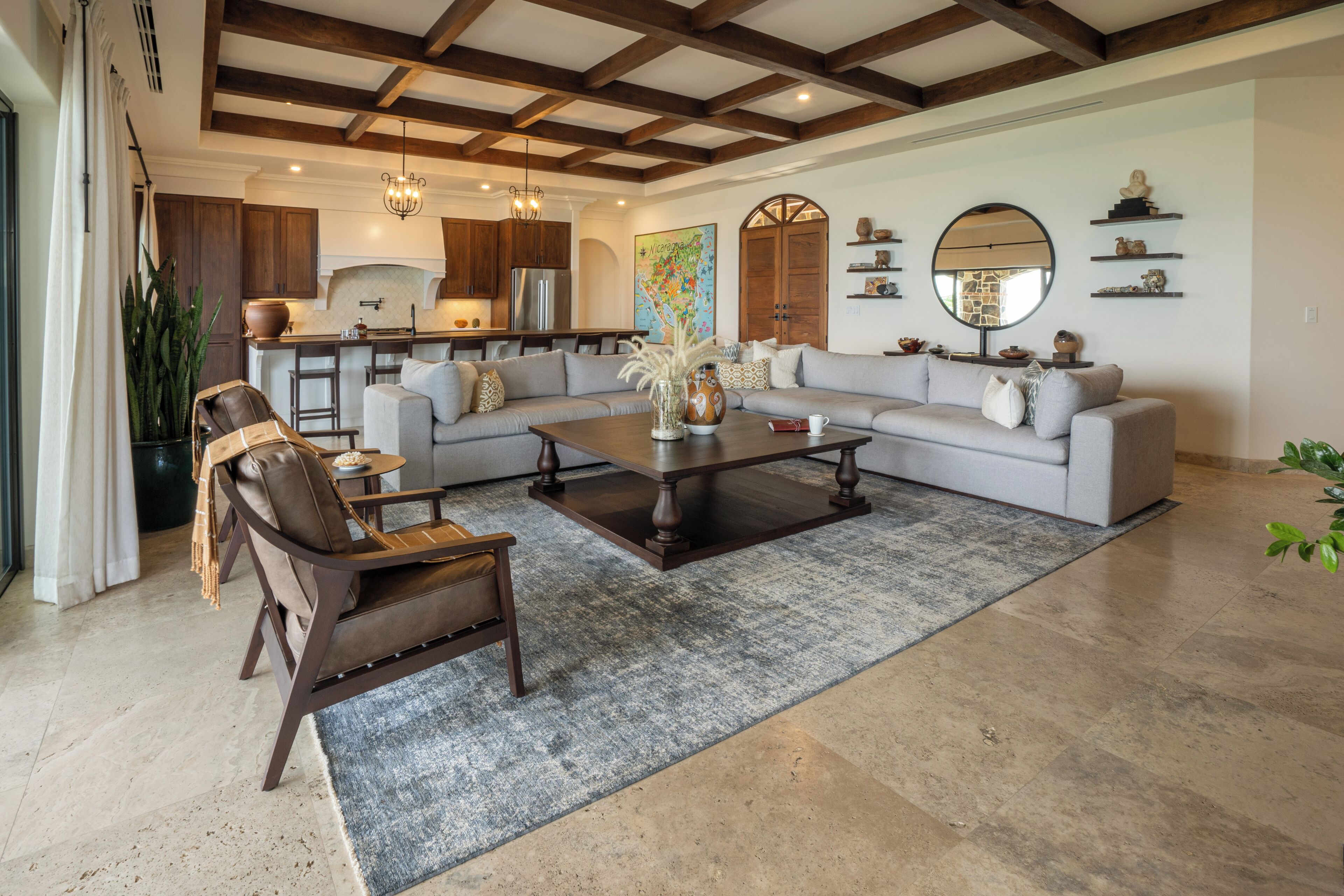 A spacious living room with a large sectional sofa, a coffee table, and a leather armchair. There is a kitchen in the background with modern appliances and wooden cabinets.
