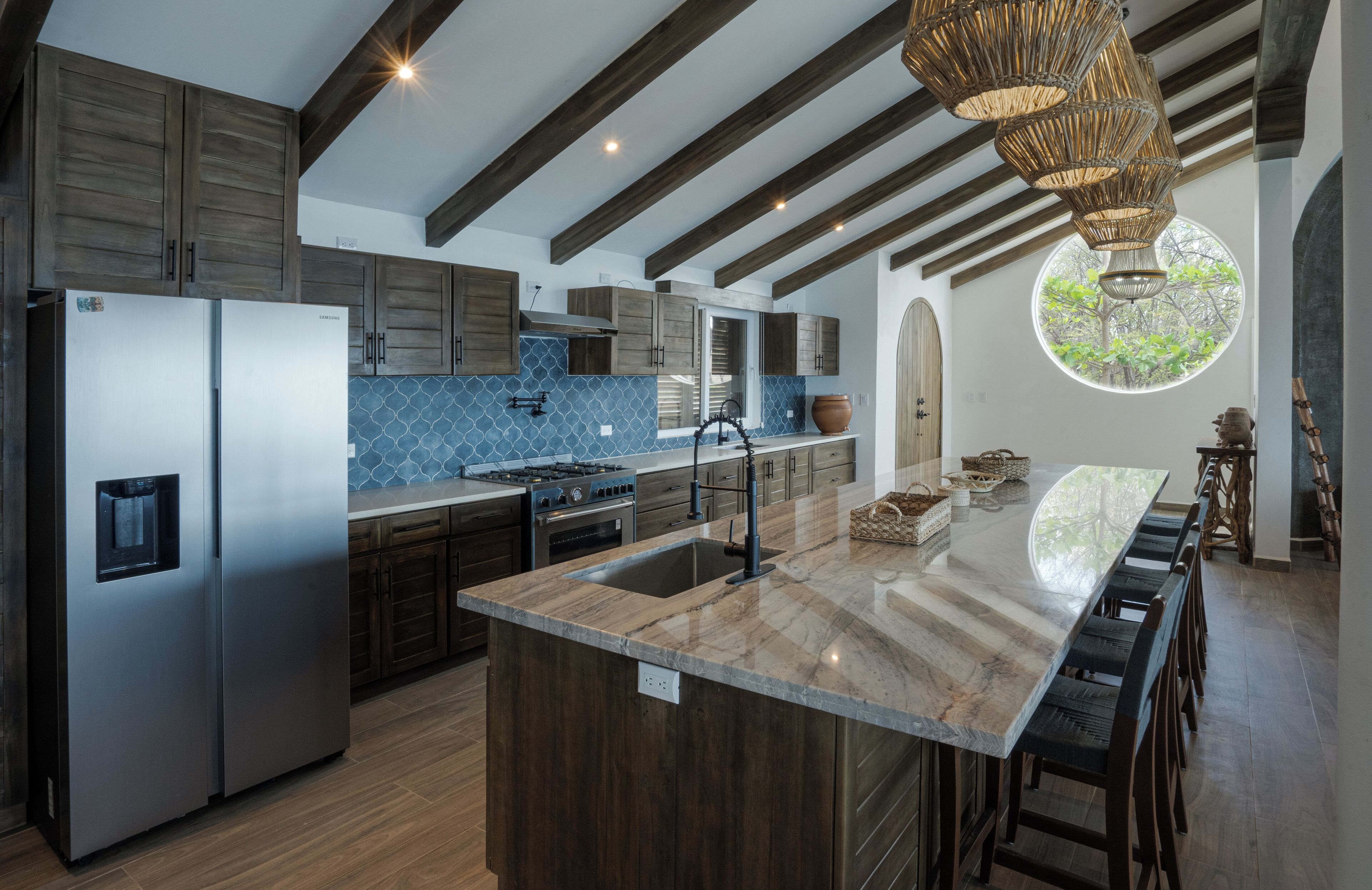 A modern kitchen with a large island, stainless steel appliances, and a view of greenery through a circular window.