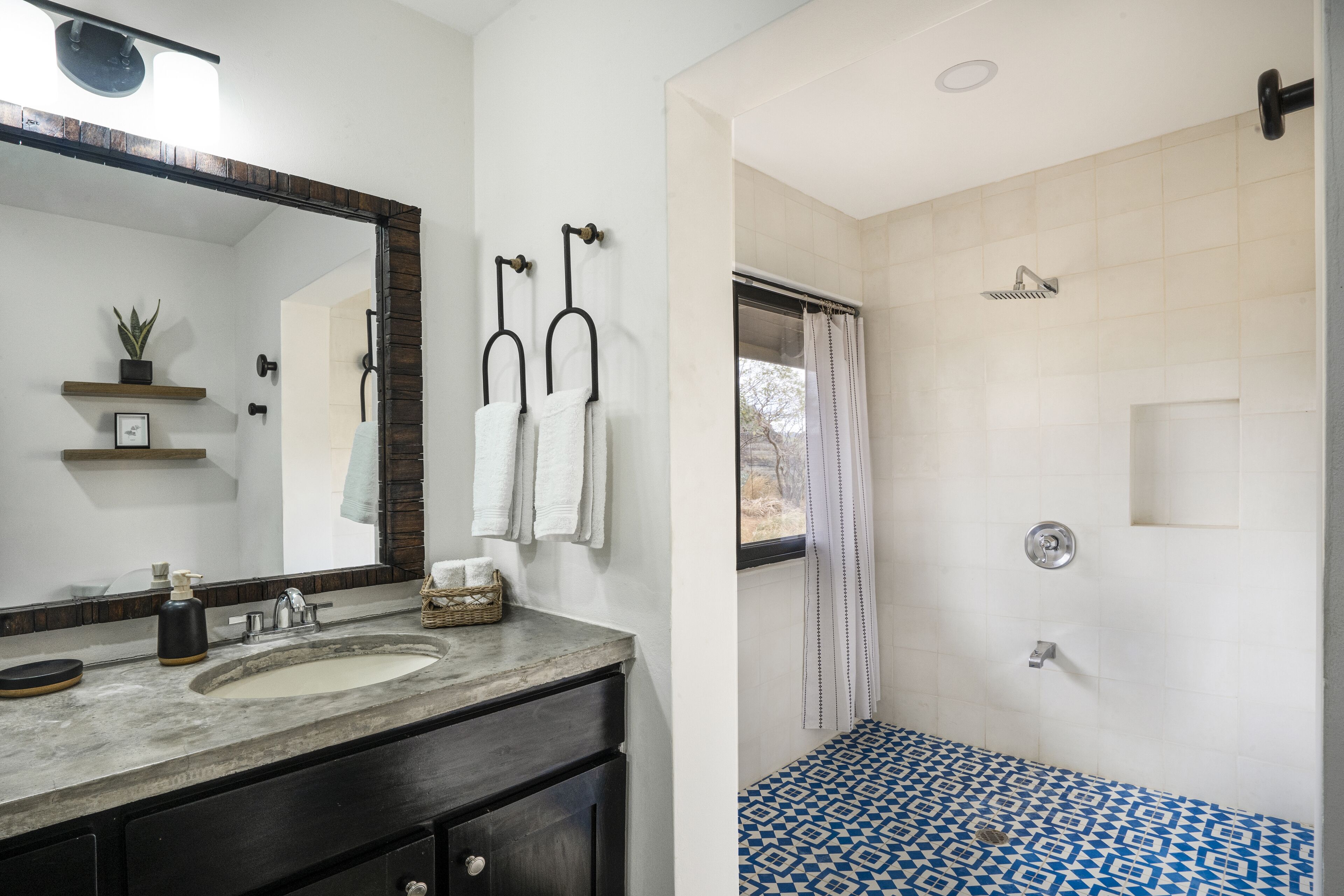 Une salle de bain avec un grand miroir, un lavabo, deux serviettes suspendues, une douche dont le sol est recouvert de carreaux bleus et blancs, et une petite fenêtre.