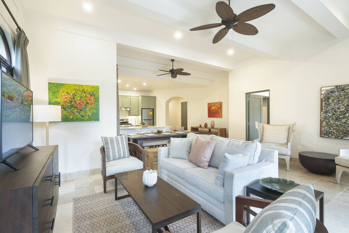 A modern living room with a sofa, coffee table, and TV. There are ceiling fans, artwork on the walls, and a kitchen in the background.