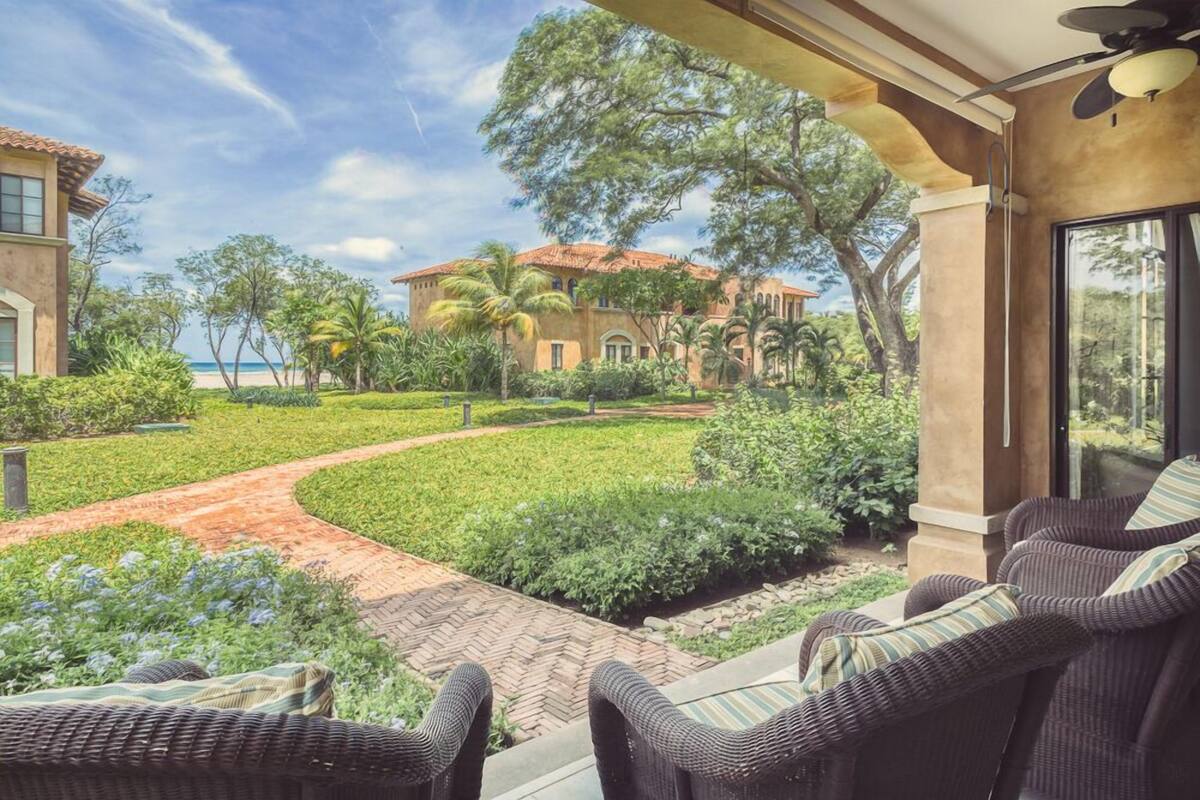 A patio with wicker furniture and a view of a landscaped garden and a building with a red roof.