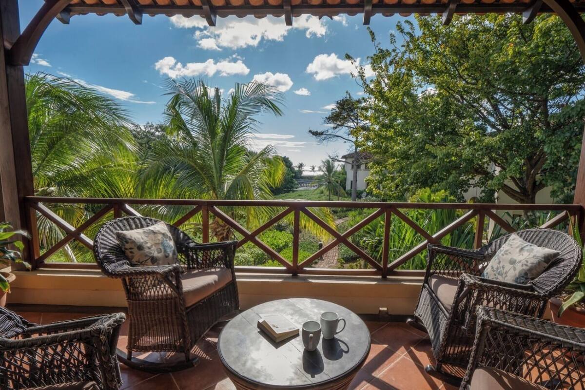 A balcony with wicker furniture, a round table with two cups and a book, and a view of lush greenery and a building in the distance.