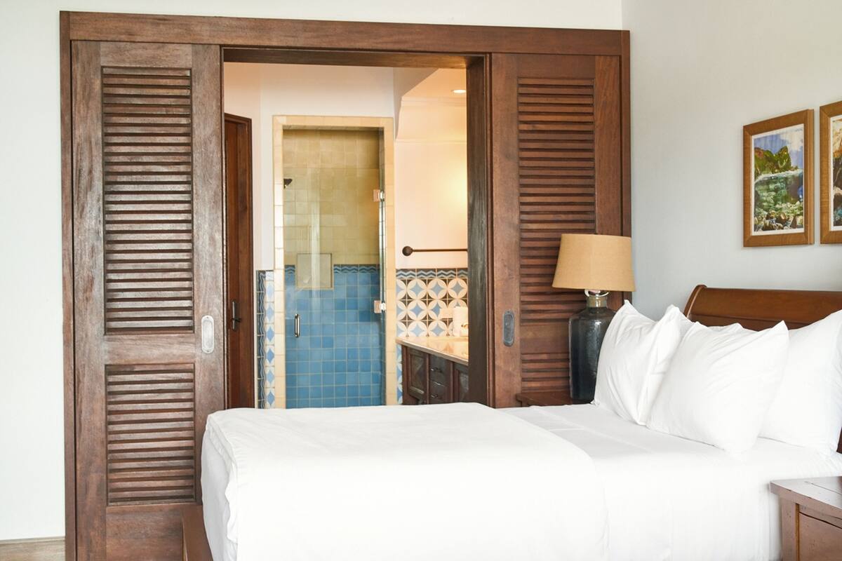 A hotel room with a bed, a nightstand, a lamp, and a bathroom visible through an open door.