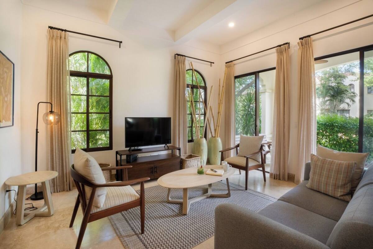 A spacious living room with a flat-screen TV, comfortable seating, and large windows offering a view of greenery.