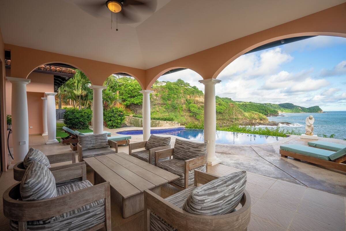 A covered outdoor seating area with a pool, white columns, and a view of the ocean and hills.