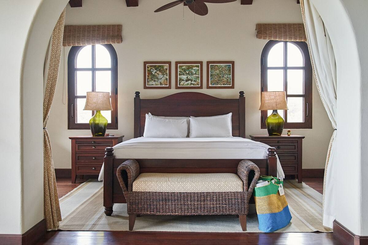 A bedroom with a wooden bed, two nightstands, a bench, and a large window with curtains.