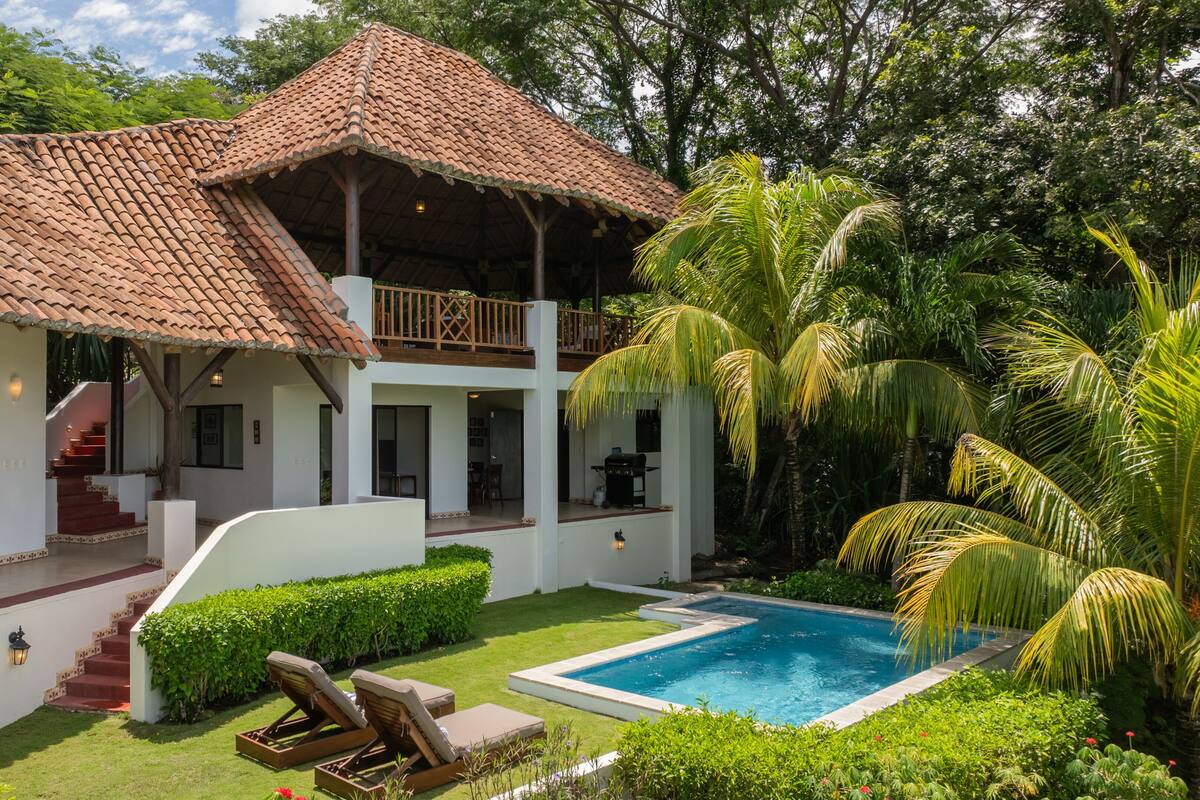 A two-story house with a tiled roof, a swimming pool, and lush greenery.