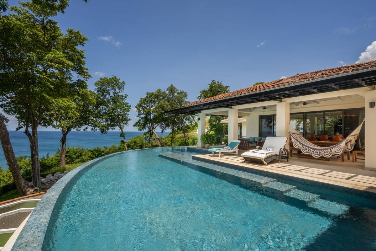 A clear blue infinity pool with lounge chairs and a hammock, overlooking a scenic ocean view.