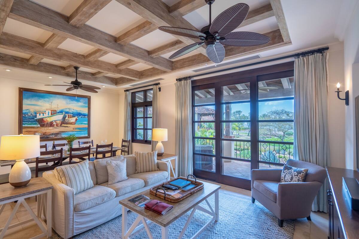 A spacious living area with a high ceiling, wooden beams, a large window with a view, a dining area with a table and chairs, and a cozy seating arrangement.