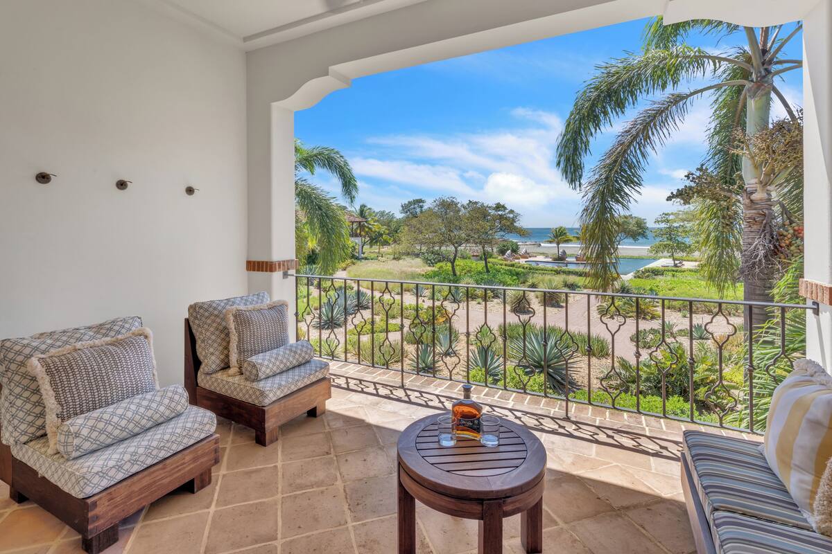 A balcony with two sofas, a small table, and a view of a landscaped garden and beach.