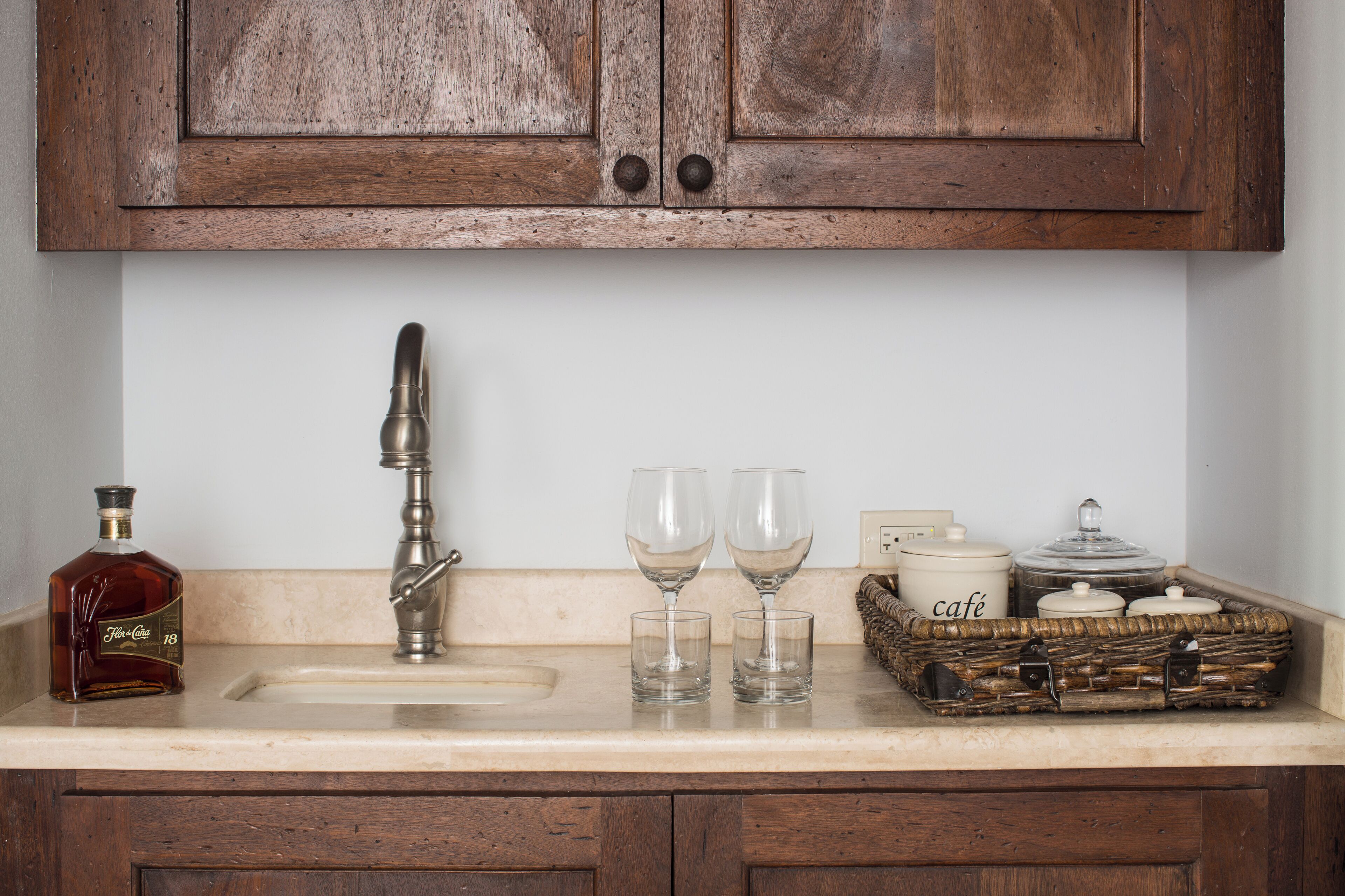 A kitchen with wooden cabinets, a sink, a bottle of liquor, two wine glasses, and a wicker basket.
