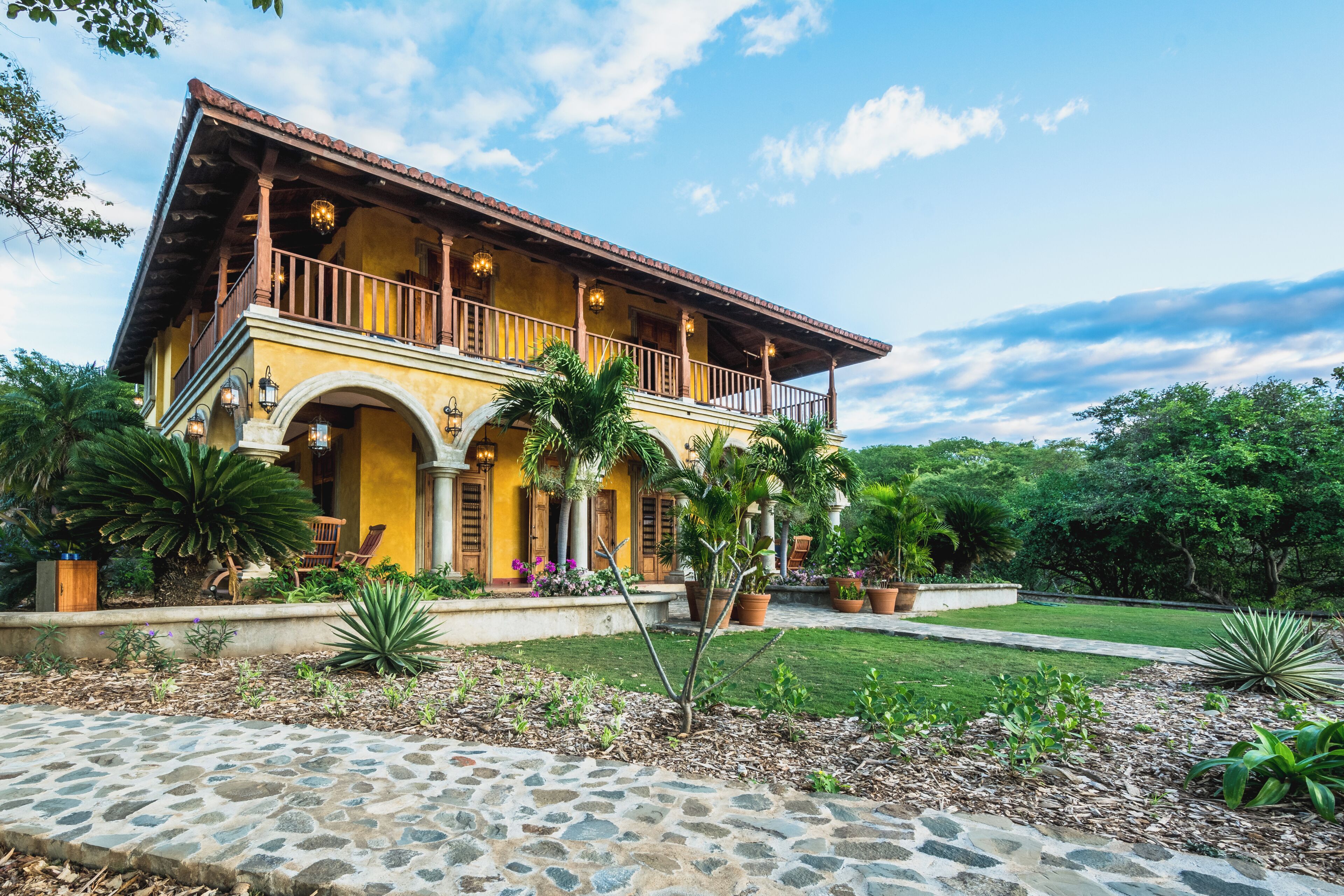 A two-story building with a balcony, stone pathway, and tropical landscaping.