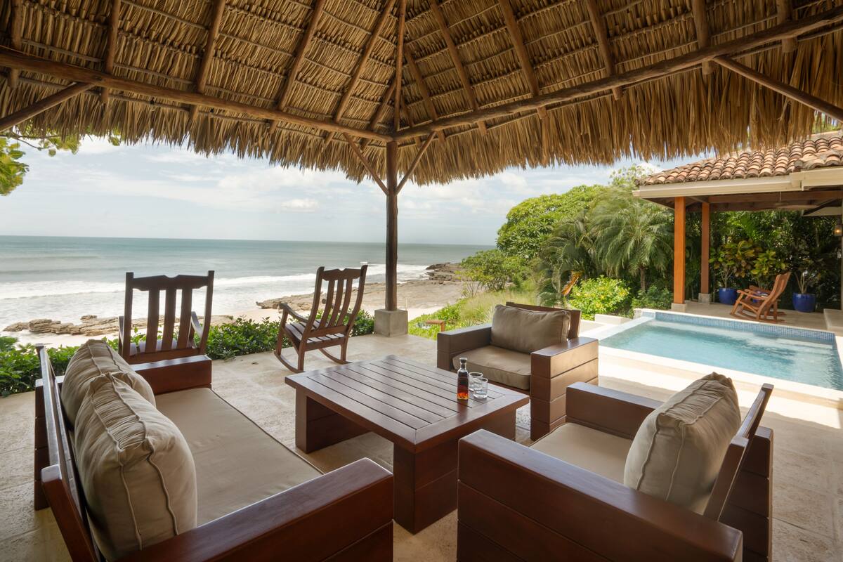 A beachside patio with a pool, wooden furniture, and a view of the ocean.