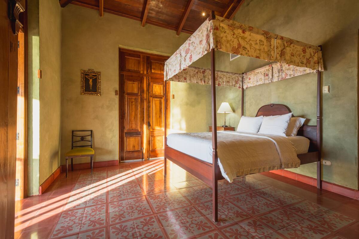 A four-poster bed with a floral canopy, a wooden chair, a wooden door, and a patterned floor.