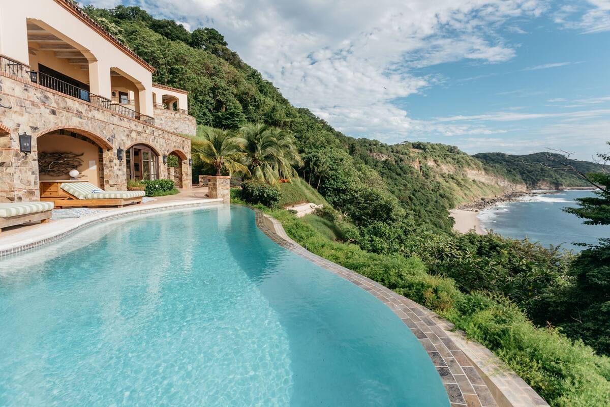 A curved infinity pool with a stone building adjacent to it, overlooking a beach and lush greenery.