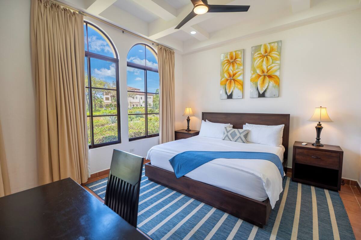 A bedroom with a large bed, two bedside tables with lamps, a chair, a desk, and a view of greenery outside.