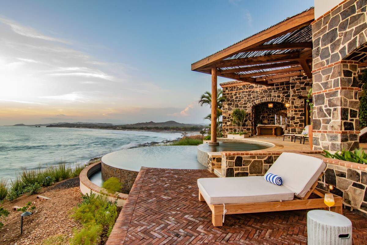 A stone building with a pool and lounge chairs overlooking the ocean.