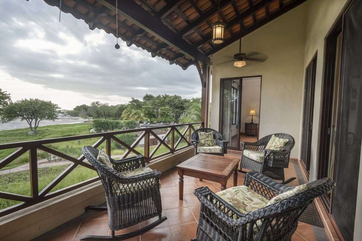 A balcony with wicker furniture, a wooden table, and a view of a river and greenery.