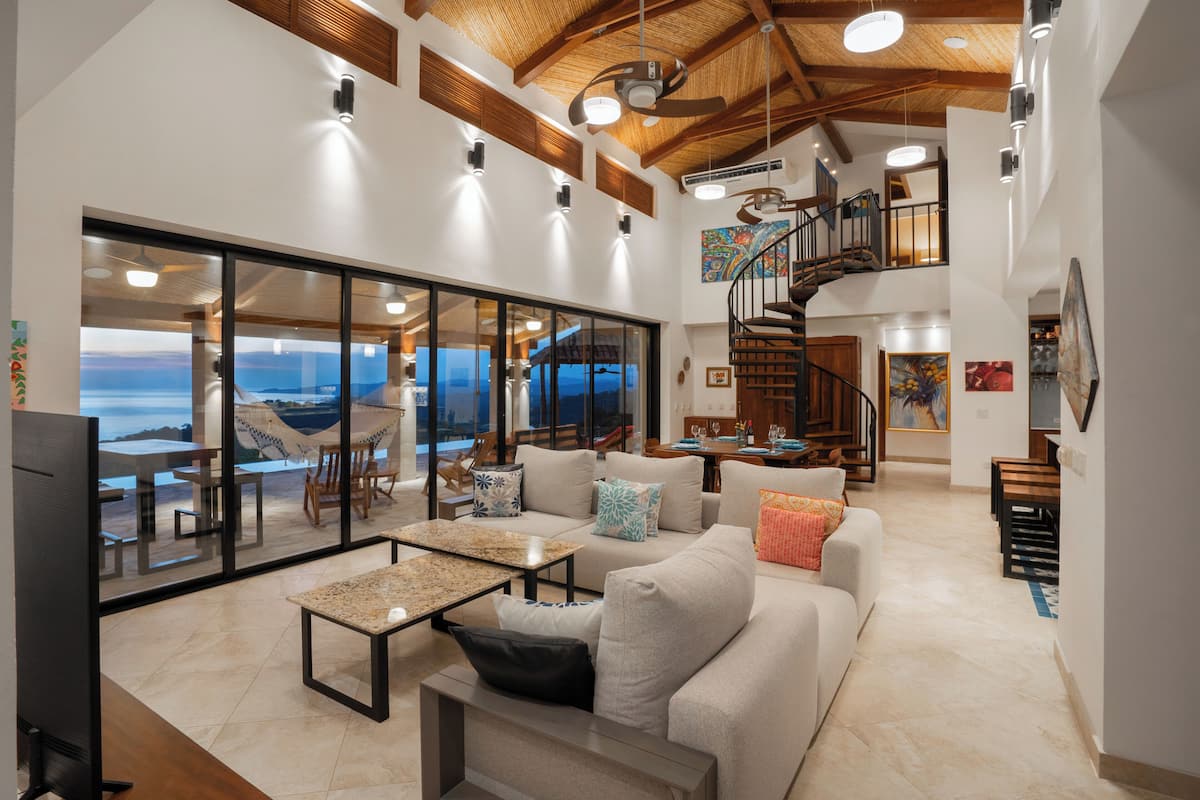 A spacious living area with a large window overlooking the sea, a comfortable sofa set, a dining area, and a staircase leading to an upper level.