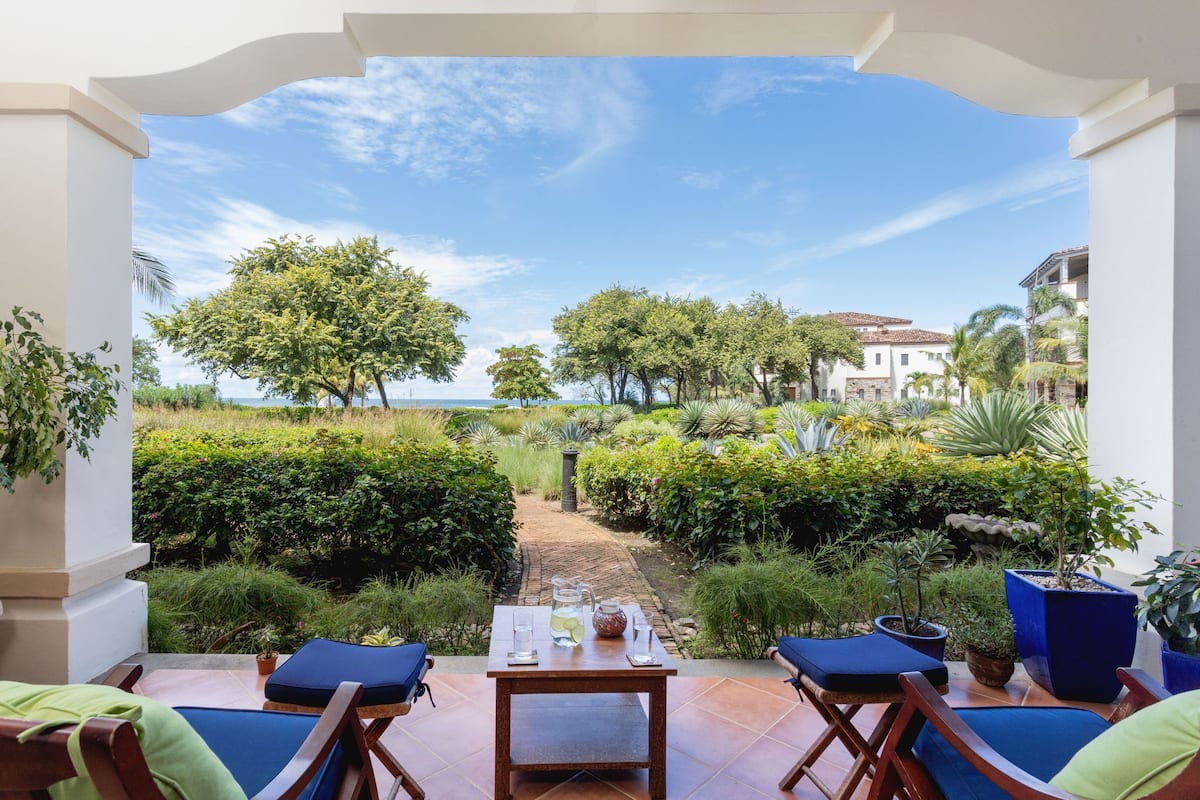 A patio with a table set for two, surrounded by lush greenery and a view of the sea.