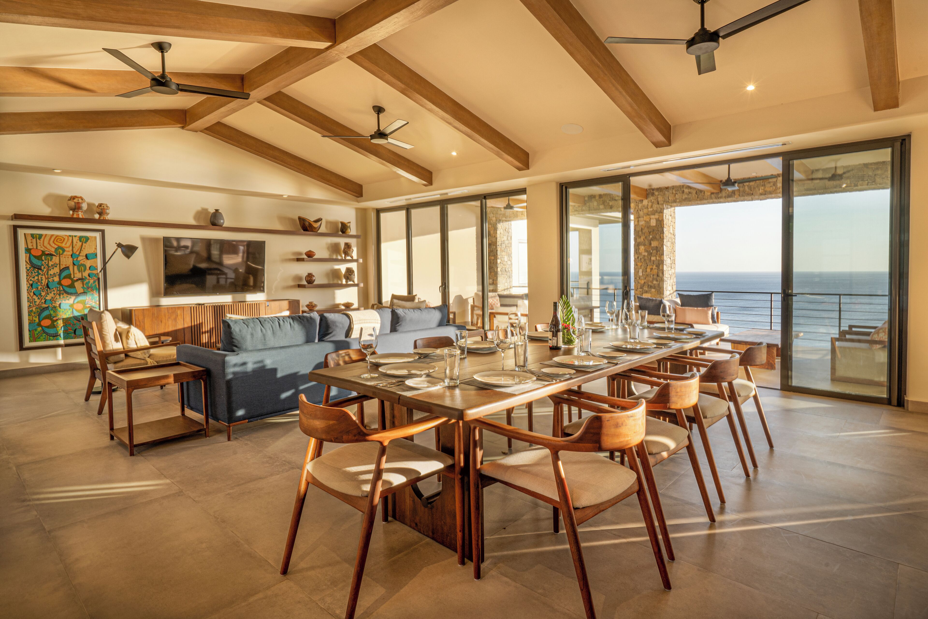 A spacious dining area with a long table, wooden chairs, and a view of the ocean through large windows.