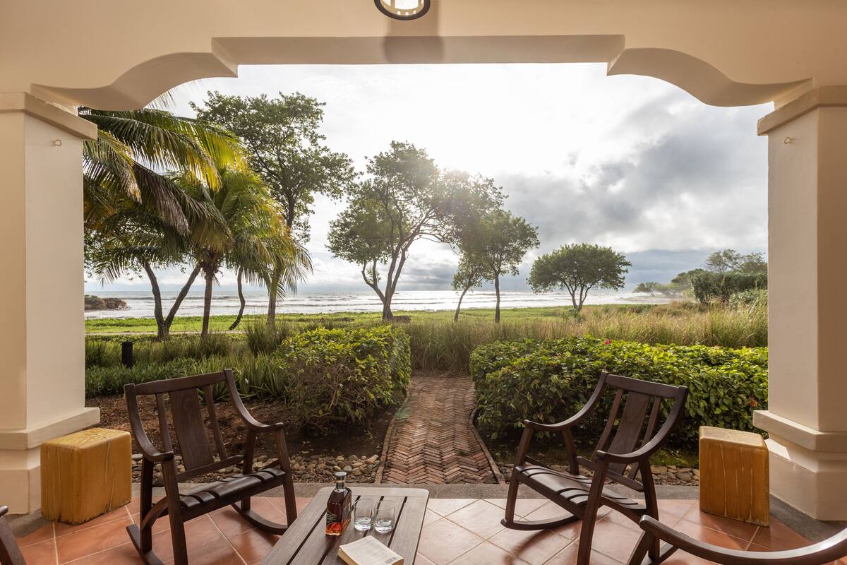 A patio with two wooden chairs and a table, overlooking a beach with palm trees and a herringbone brick pathway.