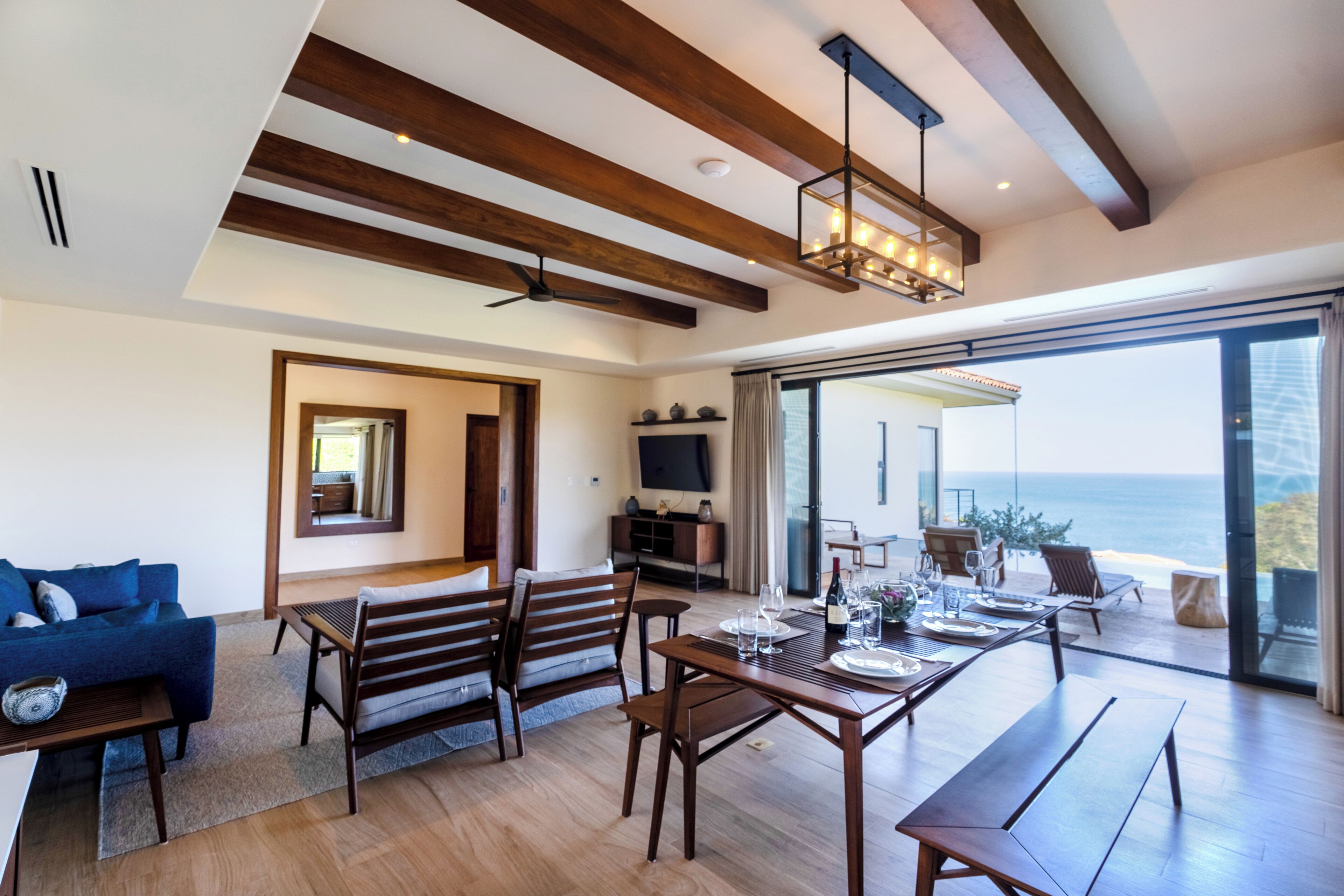 A spacious living area with a wooden dining table, benches, and a sofa. A television is mounted on the wall, and there is a view of the ocean through large glass doors.