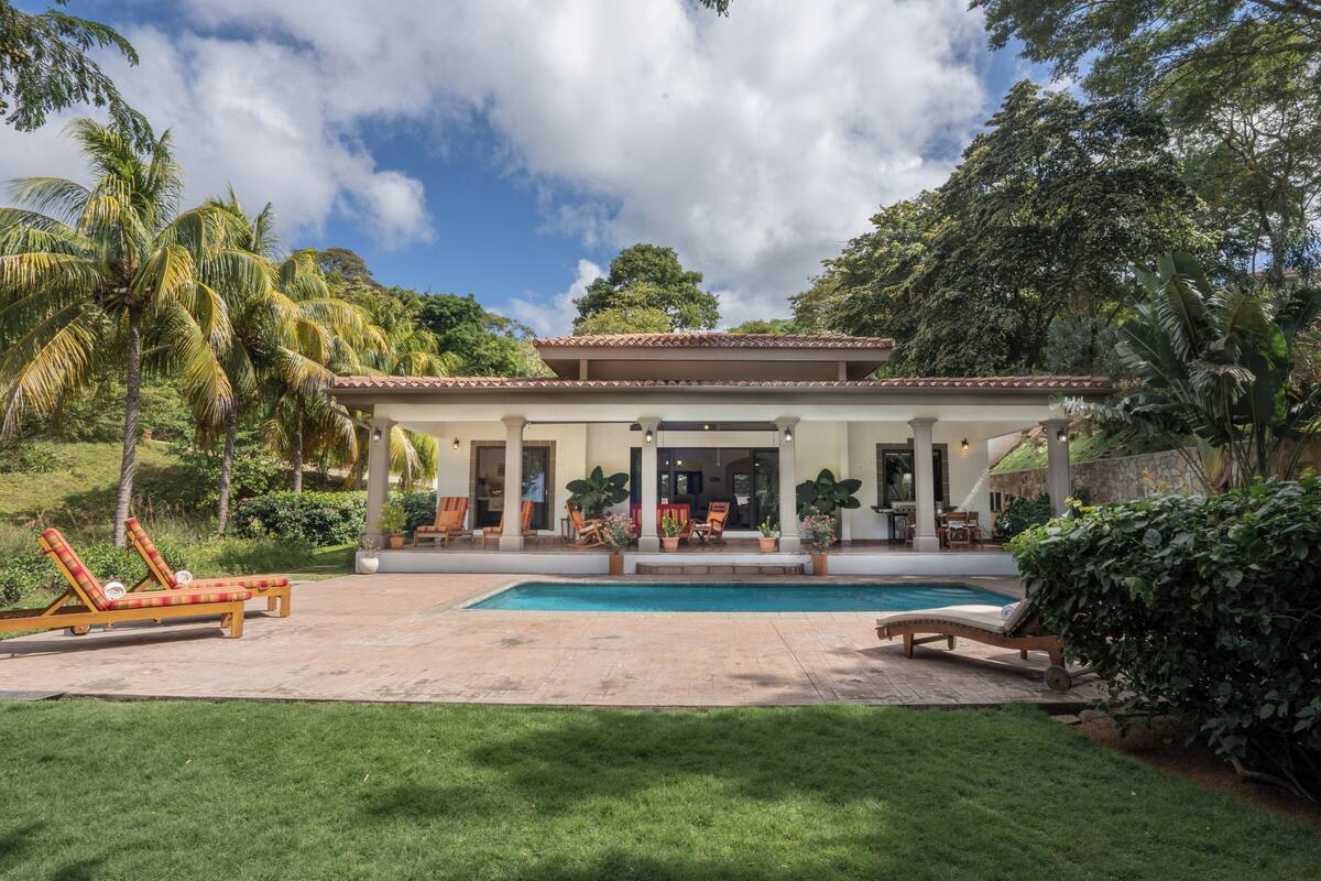 A house with a swimming pool, outdoor seating, and lush greenery.