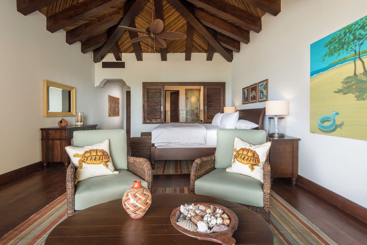 A spacious room with a large bed, two armchairs, a wooden ceiling, and a painting of a beach scene.