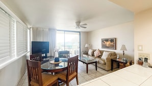 A living room with a dining table, chairs, a sofa, a TV, and a ceiling fan. - One Bedroom Ocean View Deluxe Luana Kai D201 (Kihei)