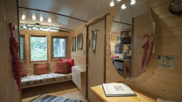 A cozy cabin interior with a wooden interior, a bed with patterned bedding, a small table with a book, and a mirror reflecting the room. - The Tiny House in Travis Heights (Austin)