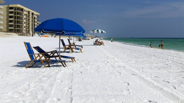 Une plage avec des transats et des parasols, un bâtiment en arrière-plan et un ciel bleu dégagé.