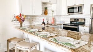 A kitchen with white cabinets, a granite countertop, a microwave, an oven, and a refrigerator. There are two bar stools and a vase with flowers on the counter. - Luana Kai Resort D306 (Kihei)