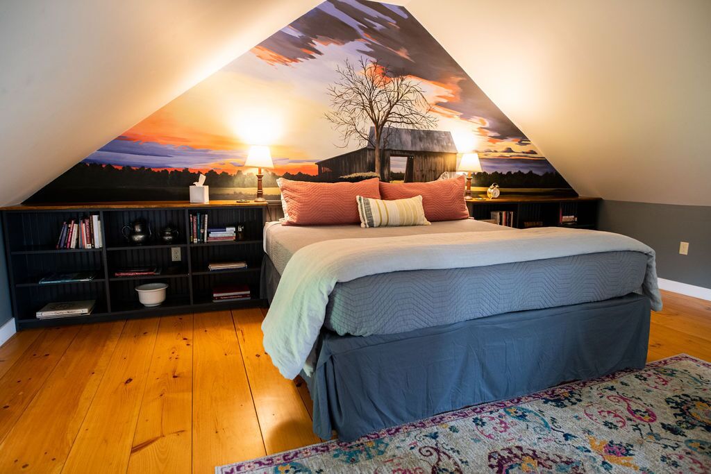 Ein Schlafzimmer mit schräg angesetzter Decke, einem Bett mit vielen Kissen, einem Bücherregal und einem Nachttisch mit Lampe.