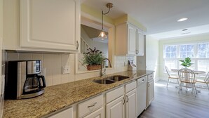 Una cocina con armarios blancos, encimera de granito, cafetera y comedor con mesa y sillas.