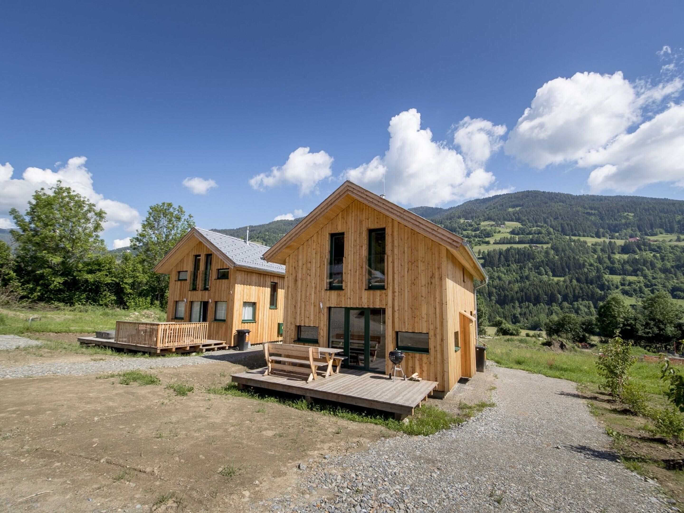 Two wooden cabins with a deck and a barbecue grill, set against a mountainous backdrop.