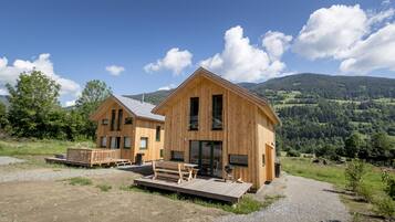 Two wooden cabins with a deck and a barbecue grill, set against a mountainous backdrop.