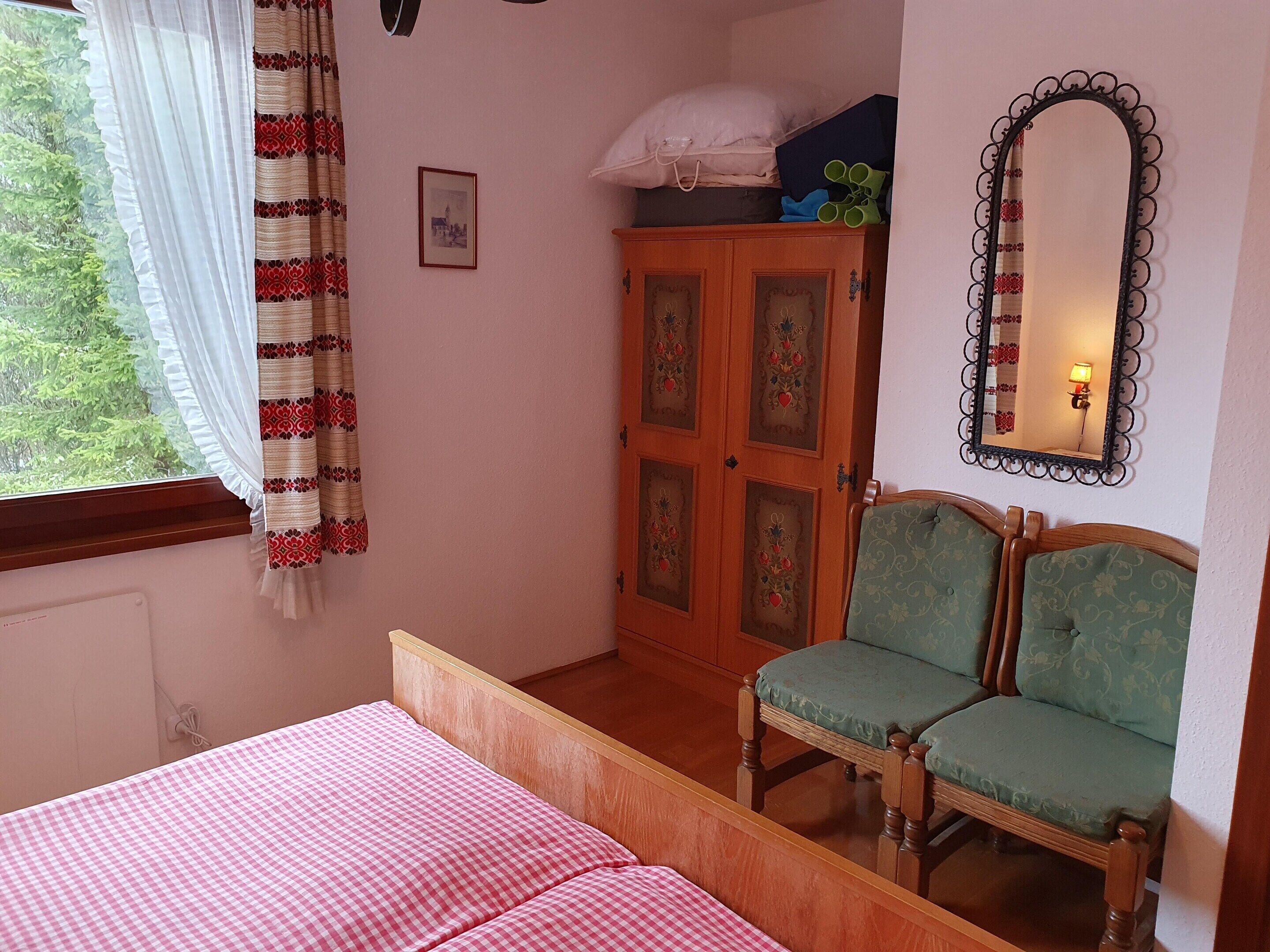 A bedroom with a bed, two chairs, a wardrobe, a mirror, and a window with curtains.