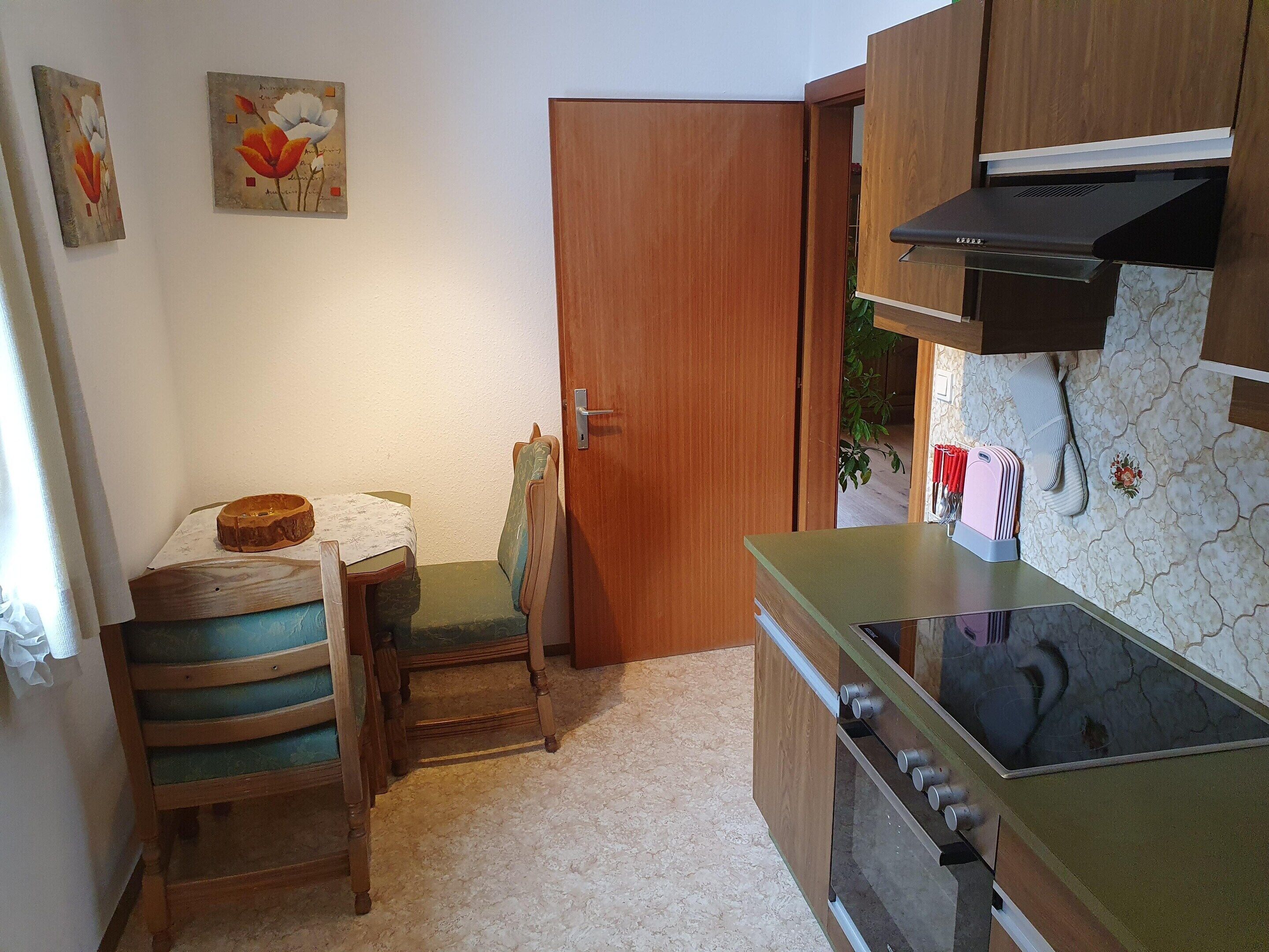A compact kitchen with wooden cabinets, a built-in oven, and a small dining area with a chair and table.